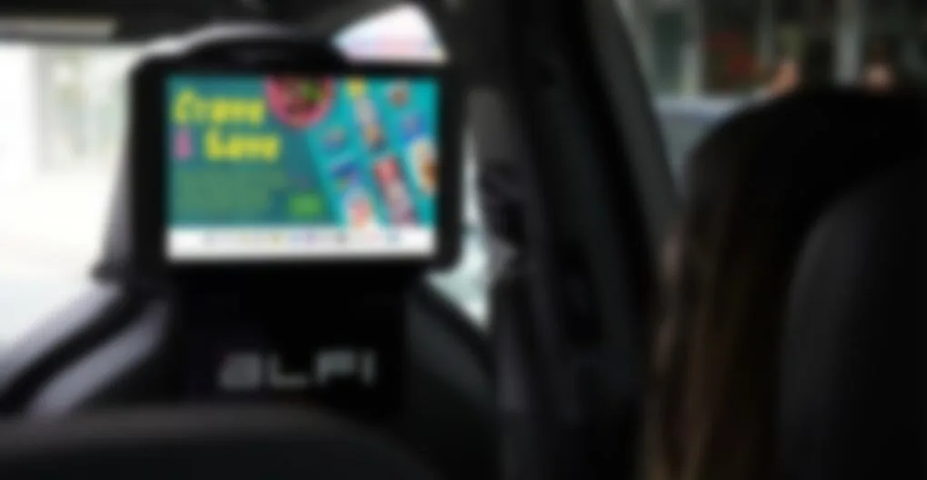 Alfi AI-enabled advertising tablet mounted on the passenger-seat headrest inside a rideshare vehicle, running a retail 'Crave & Save' campaign; ALFI branding on the mount. Photo published by DOOH trade outlet invidis.