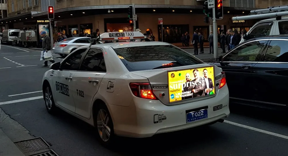 Cab Digital Media 'TaxiLive' digital rear-window billboard on a Sydney 13CABS taxi in city traffic, running a Channel Seven 'MasterChef' ('Surprises') campaign. Photo via Mediaweek.
