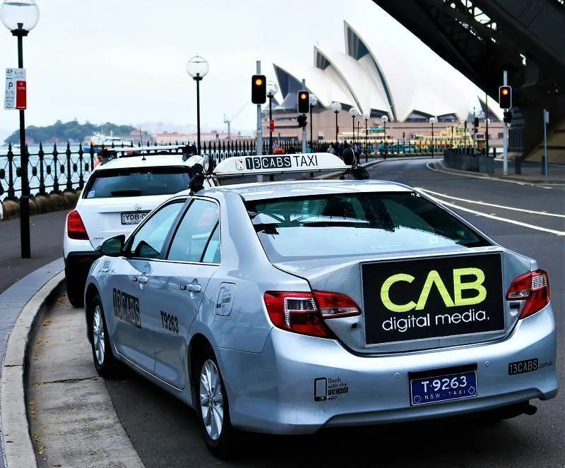 TaxiLive Solo digital billboard mounted on the rear of a 13CABS taxi (screen showing 'CAB digital media' branding) at Sydney's Circular Quay near the Opera House. High-res press photo via iothub / nextmedia.