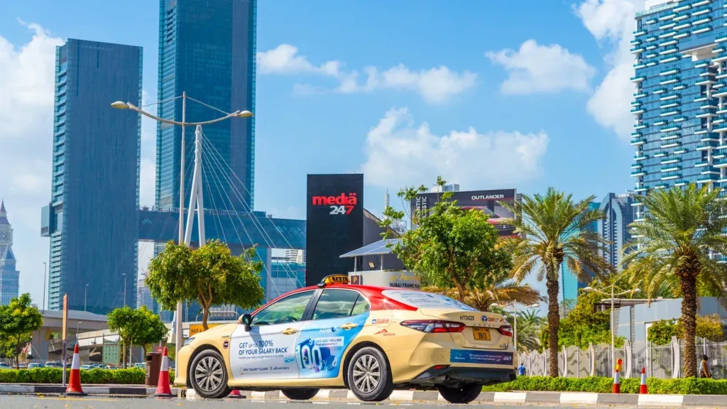 Sharaf DG 'salary back' body-wrap on a Dubai taxi, shot on Sheikh Zayed Road with a Media247 billboard behind it