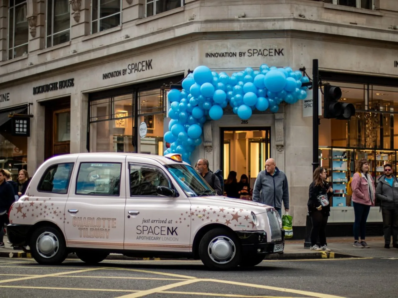 Space NK / Charlotte Tilbury wrapped taxi acting as a moving billboard on a busy shopping street with pedestrians (The Ark Marketing & Media own work)
