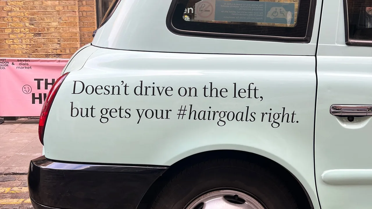 White cab superside/full-wrap reading "Doesn't drive on the left, but gets your #hairgoals right" (Transport Media taxi advertising gallery)